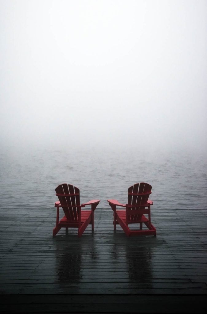 Two red adirondack chairs on a dock in the fog.