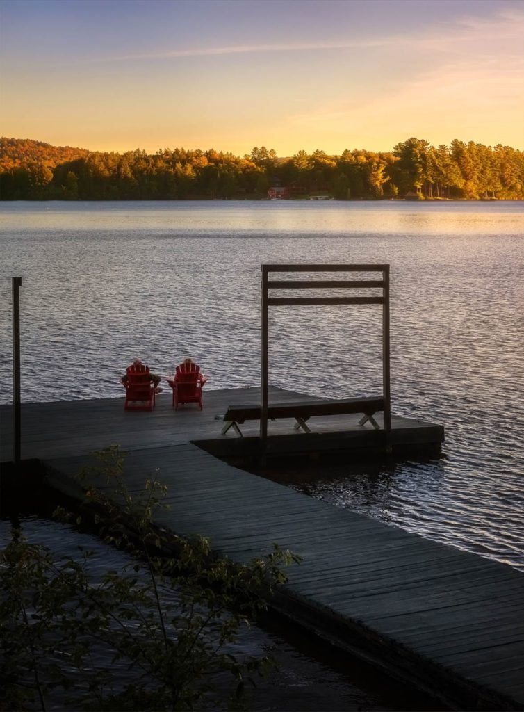 Two red chairs sit on a dock overlooking a lake.