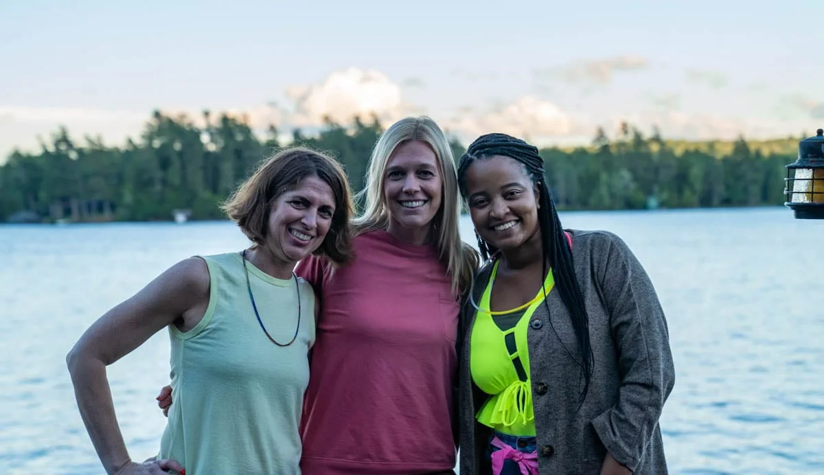 Three women posing for a photo in front of a lake.