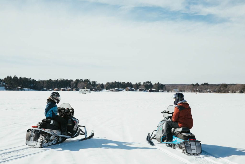Two people riding snowmobiles on a snowy field.