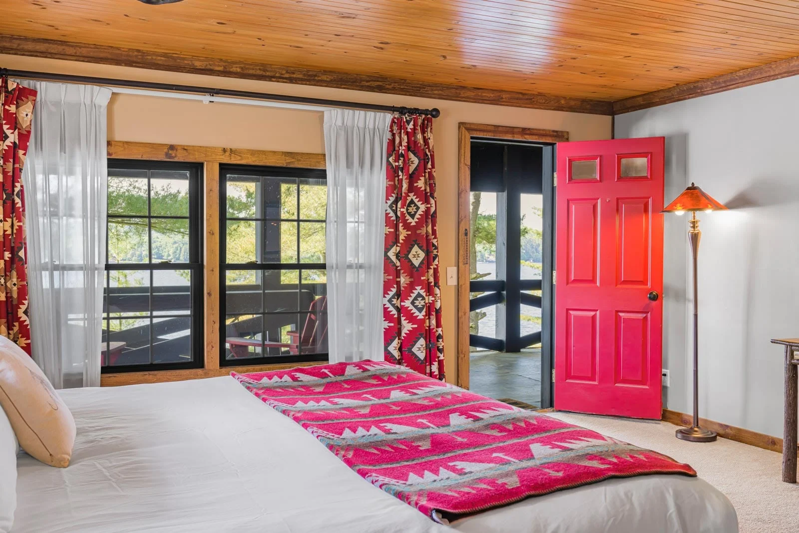 A bedroom with a wooden ceiling, a bed featuring a patterned red blanket, red curtains, a red door, and a window with a view of a wooden deck.