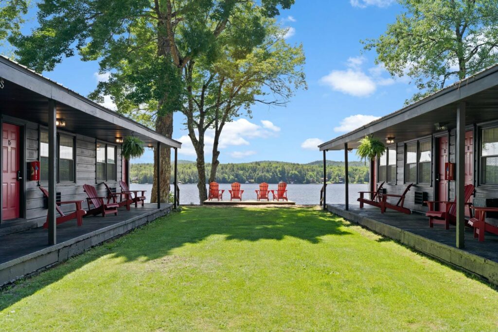 Two rows of cabins with red doors flank a grassy path leading to a lake. At the lakefront, red Adirondack chairs are lined up, offering a view of the water and distant trees.
