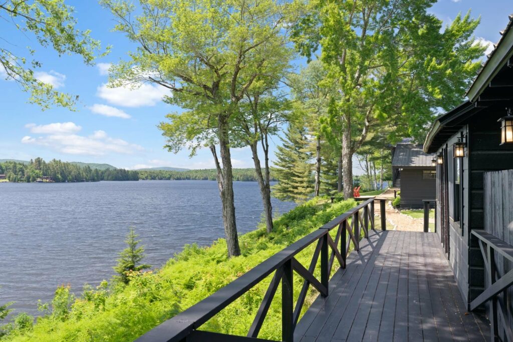 Wooden deck of a cabin overlooking a calm lake surrounded by lush trees under a clear blue sky.