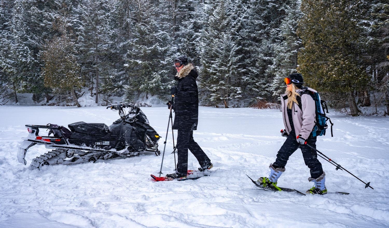 Two people snowshoeing near a snowmobile in a snowy forest.