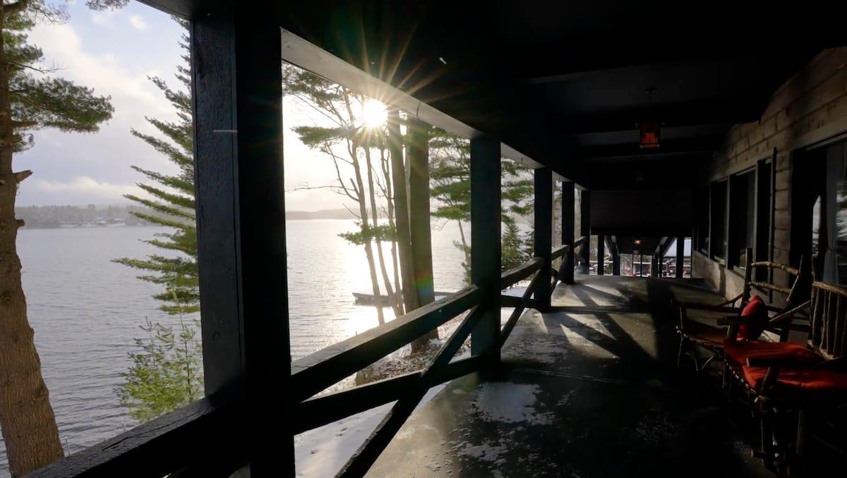 Sunset view from a wooden porch overlooking a lake, with pine trees and chairs in the foreground.