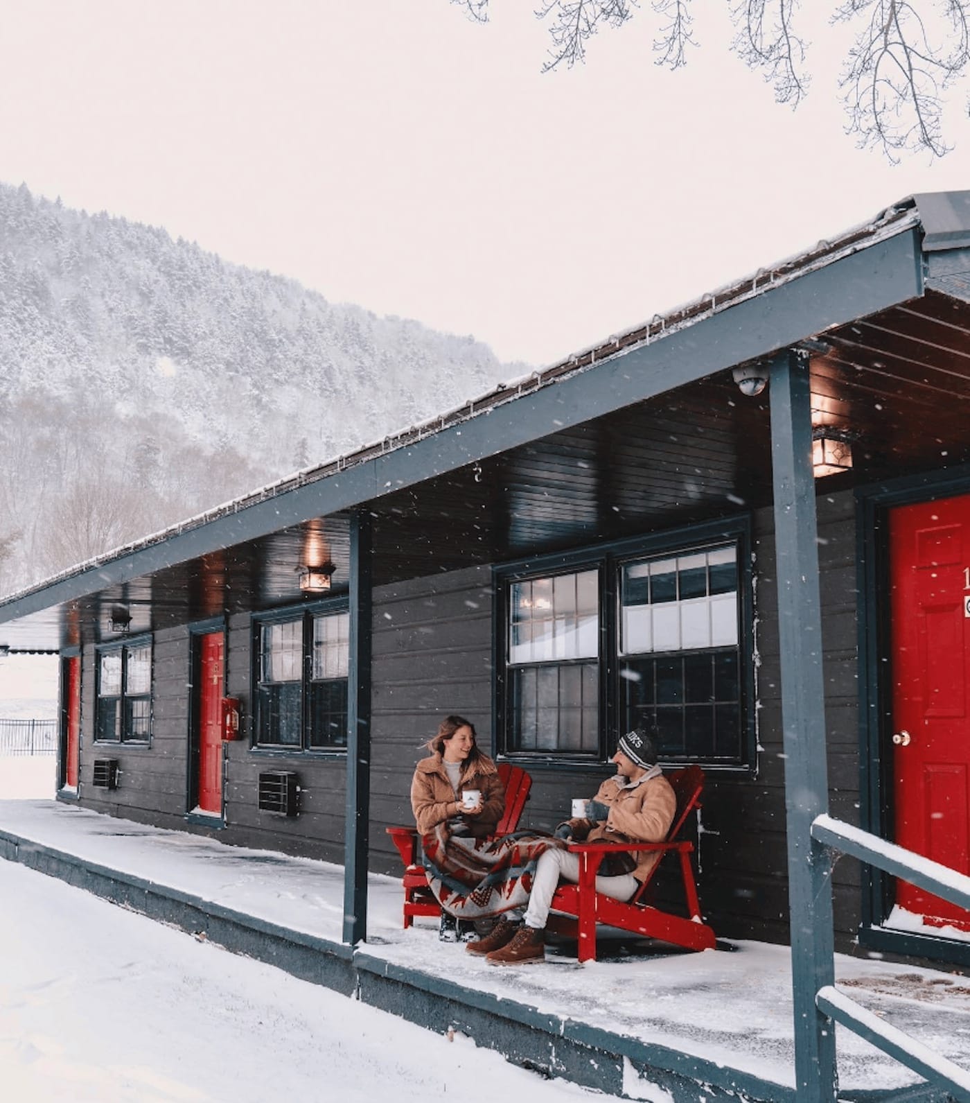 Two people sit on red chairs on a snowy porch, holding cups. The building is dark with red doors, set in a snowy mountain landscape.
