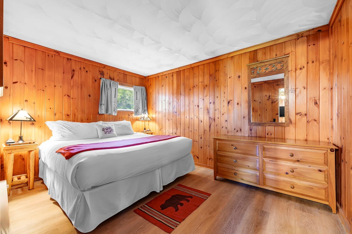 Wood-paneled bedroom with a large bed, side tables, a dresser, and a mirror. There's a small window with curtains, and a red rug is placed on the wooden floor.