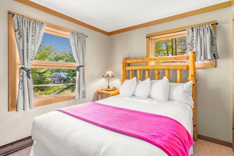 A rustic bedroom with a wooden bed frame, white linens, and a pink blanket. Two windows with patterned curtains show a view of trees and a parked car. A small lamp is on the bedside table.