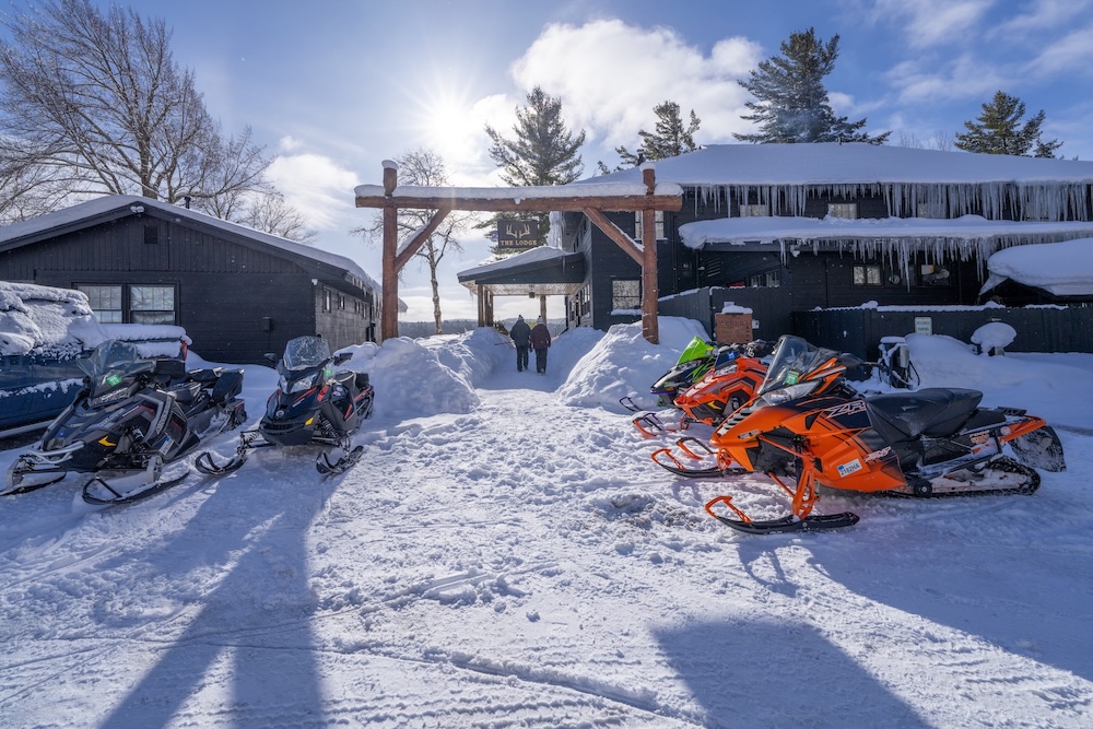 Snowmobiles parked in front of snowy lodge buildings under a sunny, clear sky, with two people walking in the distance.