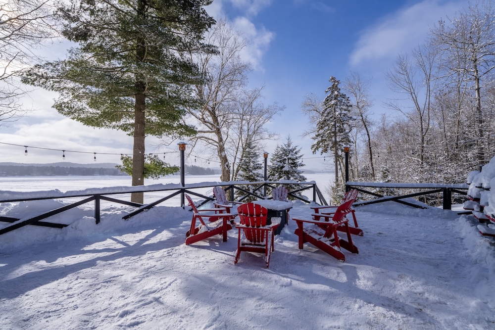 Five red wooden chairs surround a small table on a snow-covered deck, overlooking a snowy landscape with trees and a frozen lake under a clear blue sky.