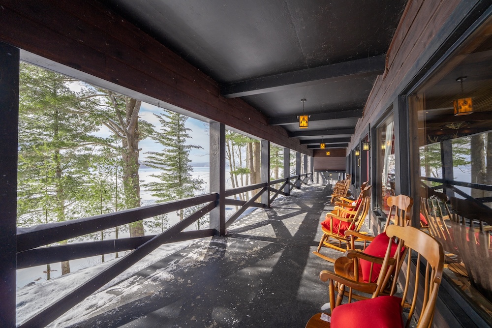 Covered porch with rocking chairs overlooking snowy landscape with trees. Snow on wooden floor, glass doors on the right, and overhead lights on a dark ceiling.