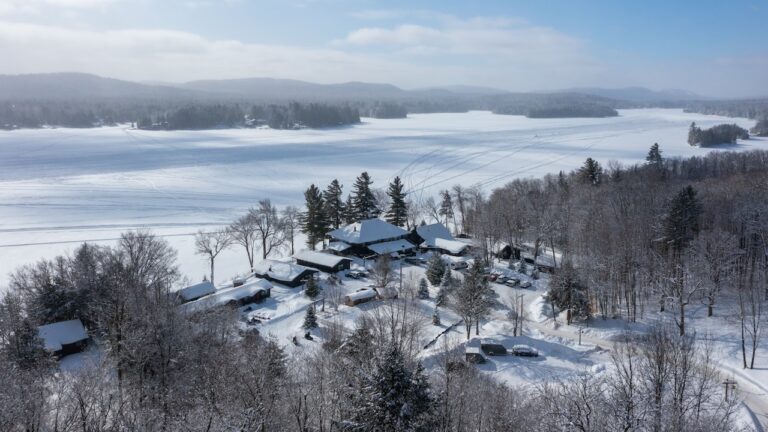 Aerial view of a snow-covered village by a frozen lake, surrounded by leafless trees and distant hills under a cloudy sky.