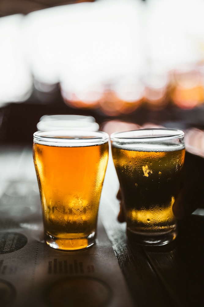 Two pint glasses filled with amber and dark beer sit on a wooden table in a dimly lit setting, with a blurred background.