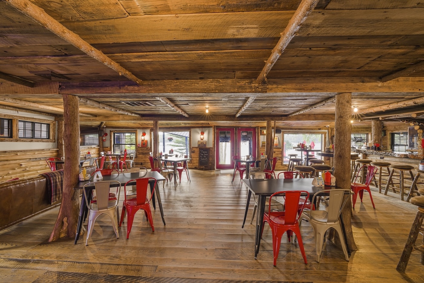 Rustic restaurant interior with wooden floors, exposed beams, metal chairs around tables, large windows, and red double doors at the back.