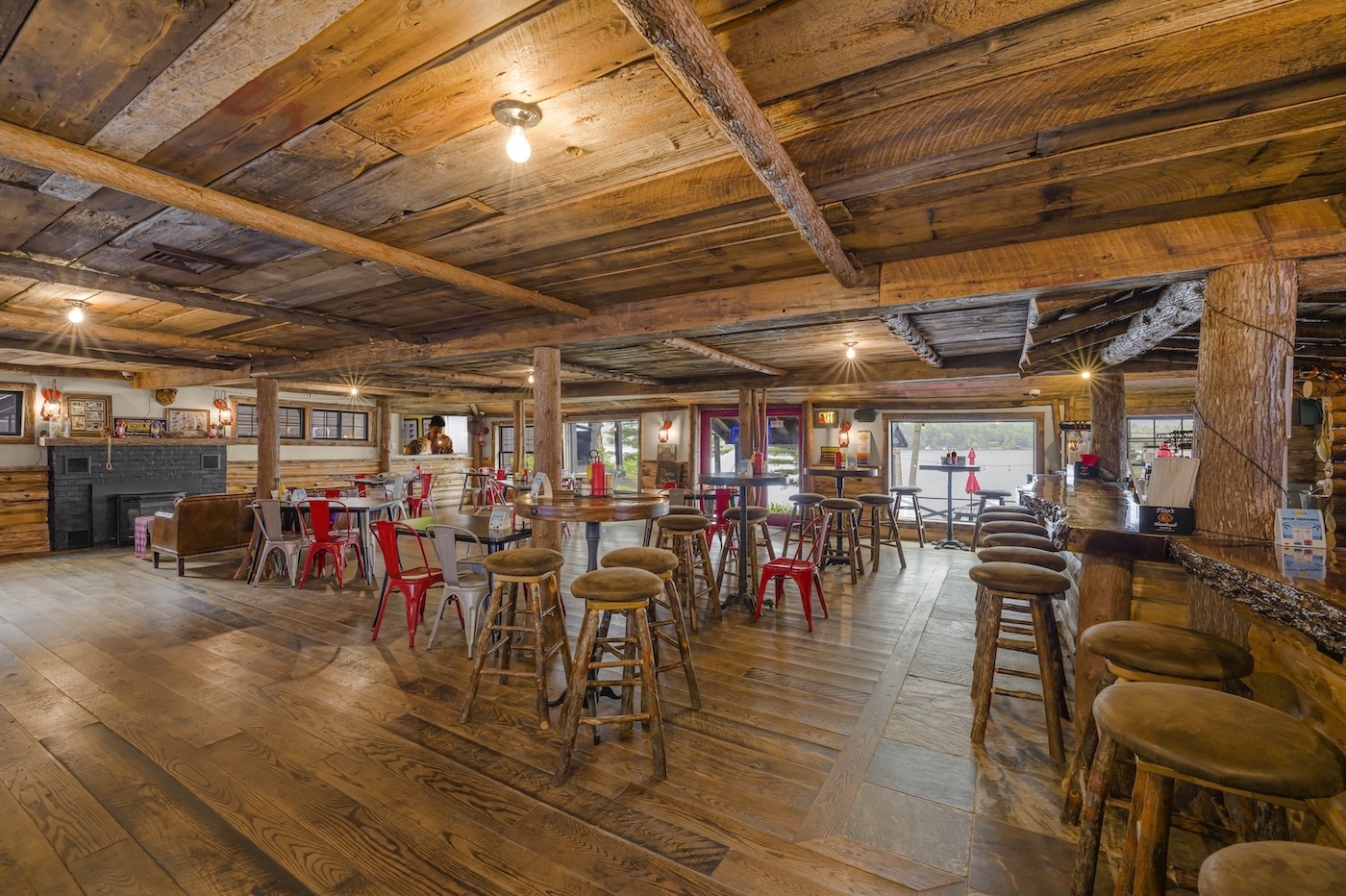 Spacious rustic interior with wooden floors and ceiling, round tables, bar stools, red chairs, and large windows overlooking an outdoor area.