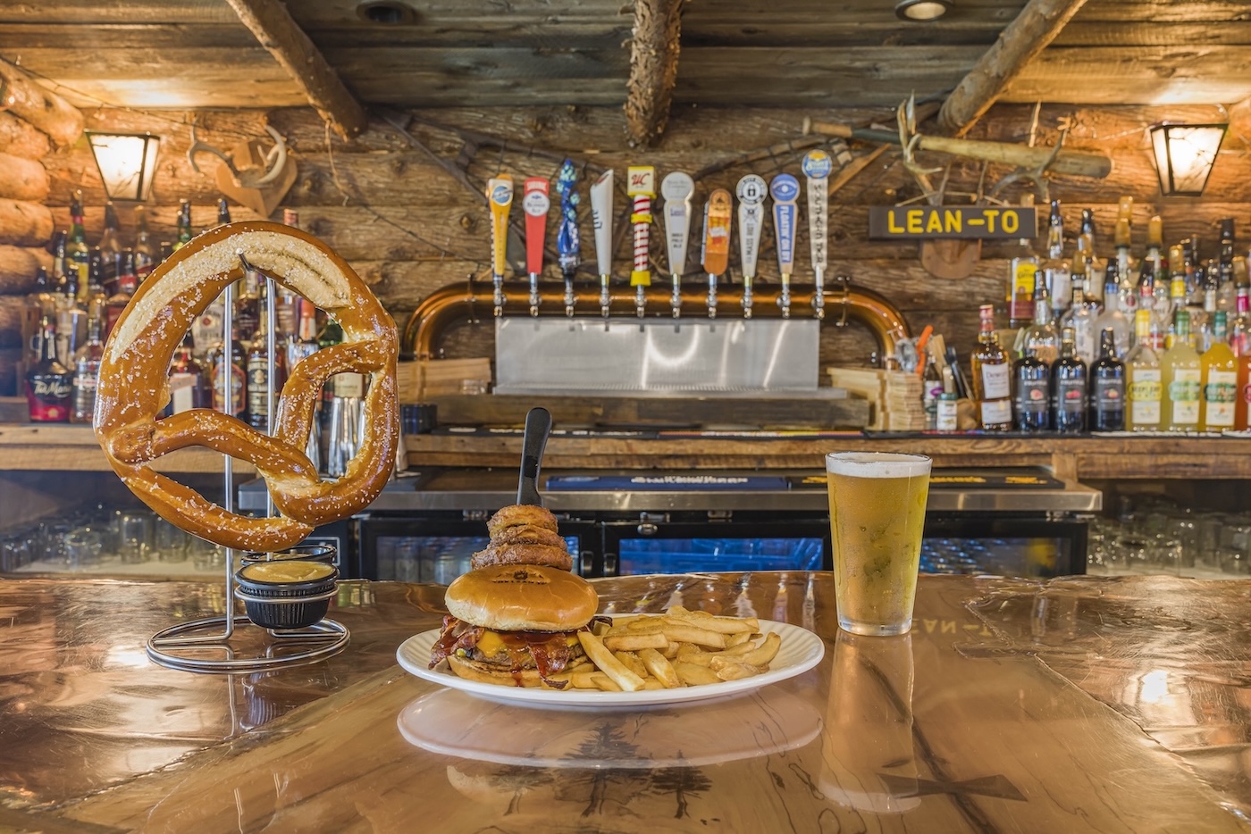 A large pretzel, a burger with fries, and a pint of beer on a wooden bar counter with beer taps and liquor bottles in the background.