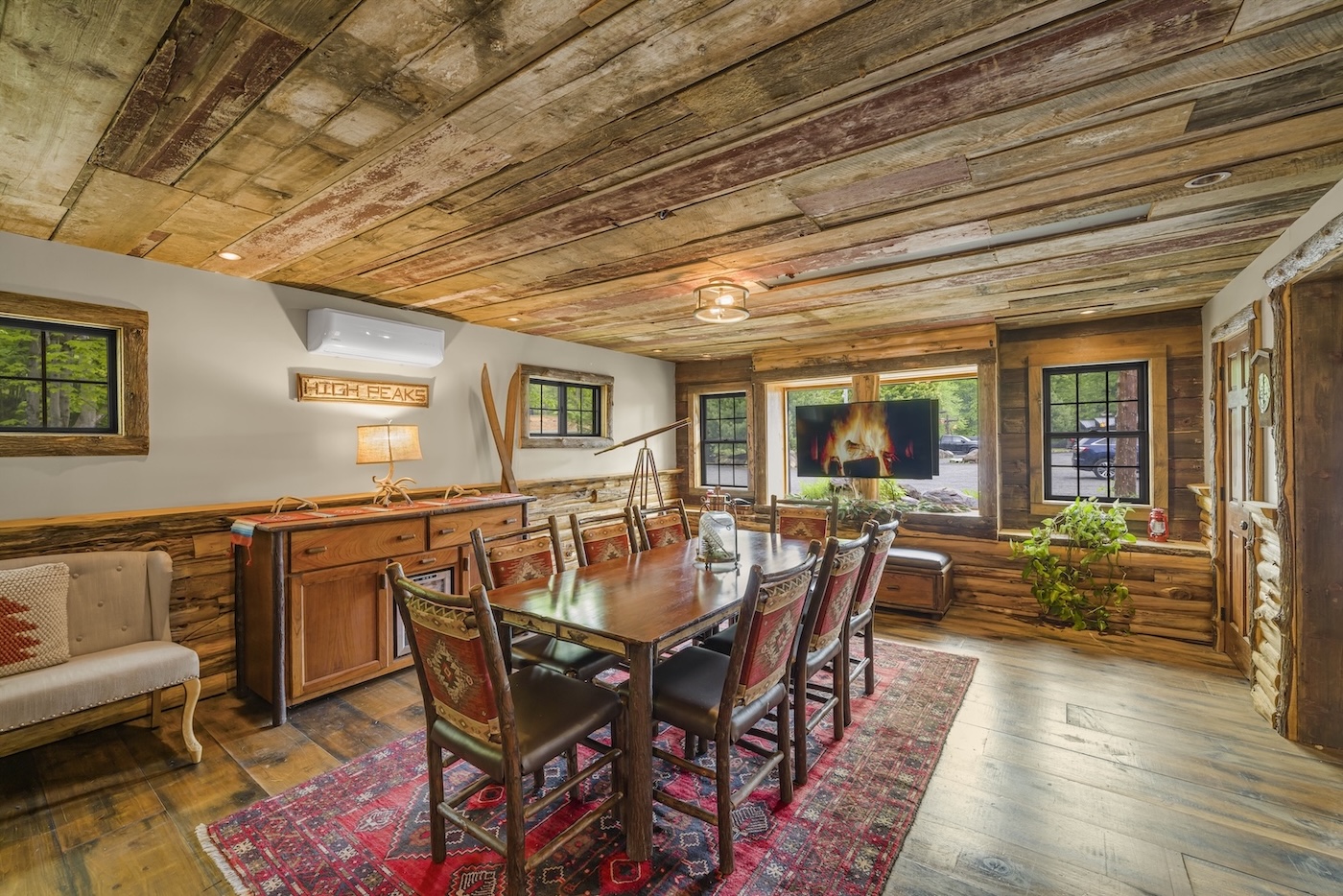 Rustic dining room with wooden walls and ceiling, a long table with eight chairs, bench seating by windows, a TV displaying a fireplace, and a red patterned rug on wood flooring.