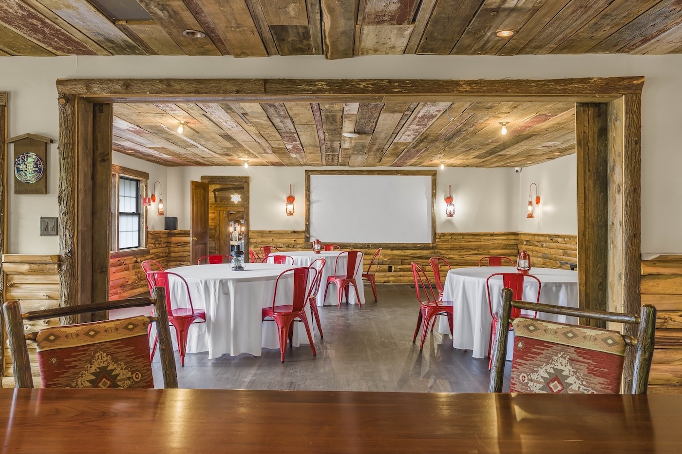 A rustic dining room with wooden walls and ceiling, round tables covered with white tablecloths, red metal chairs, and vintage-style decor.
