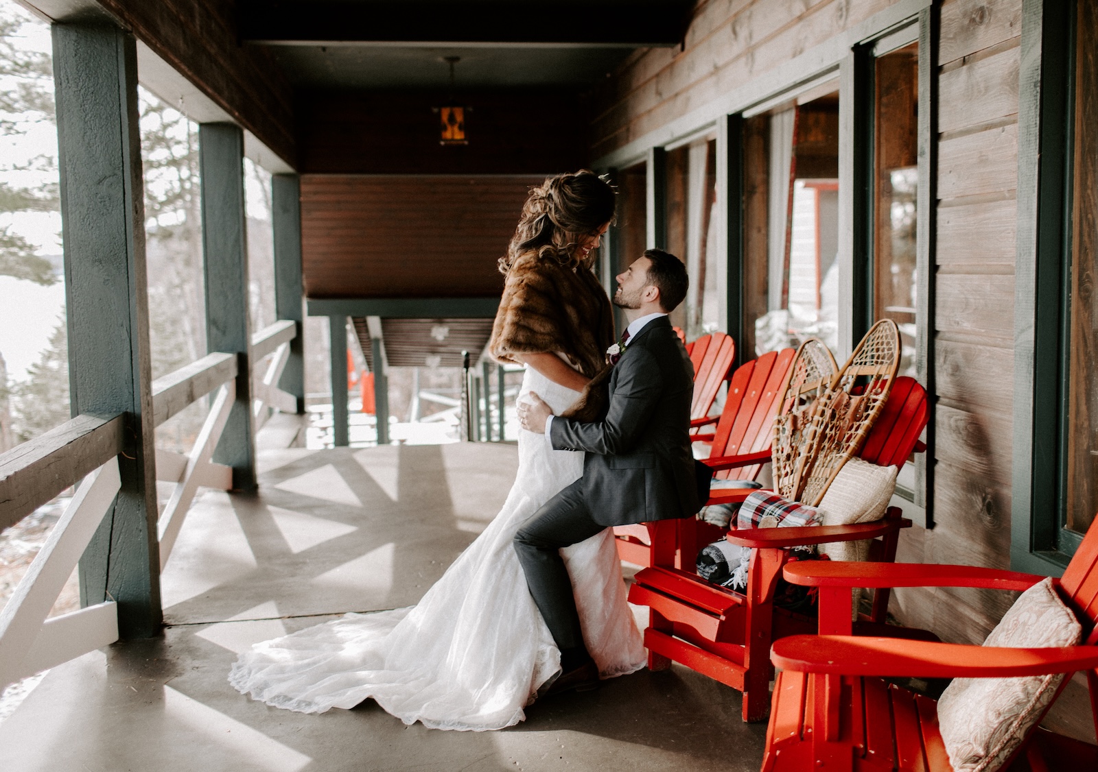 A bride and groom pose together on a rustic porch with red Adirondack chairs, the bride in a white dress and fur stole, the groom in a suit.