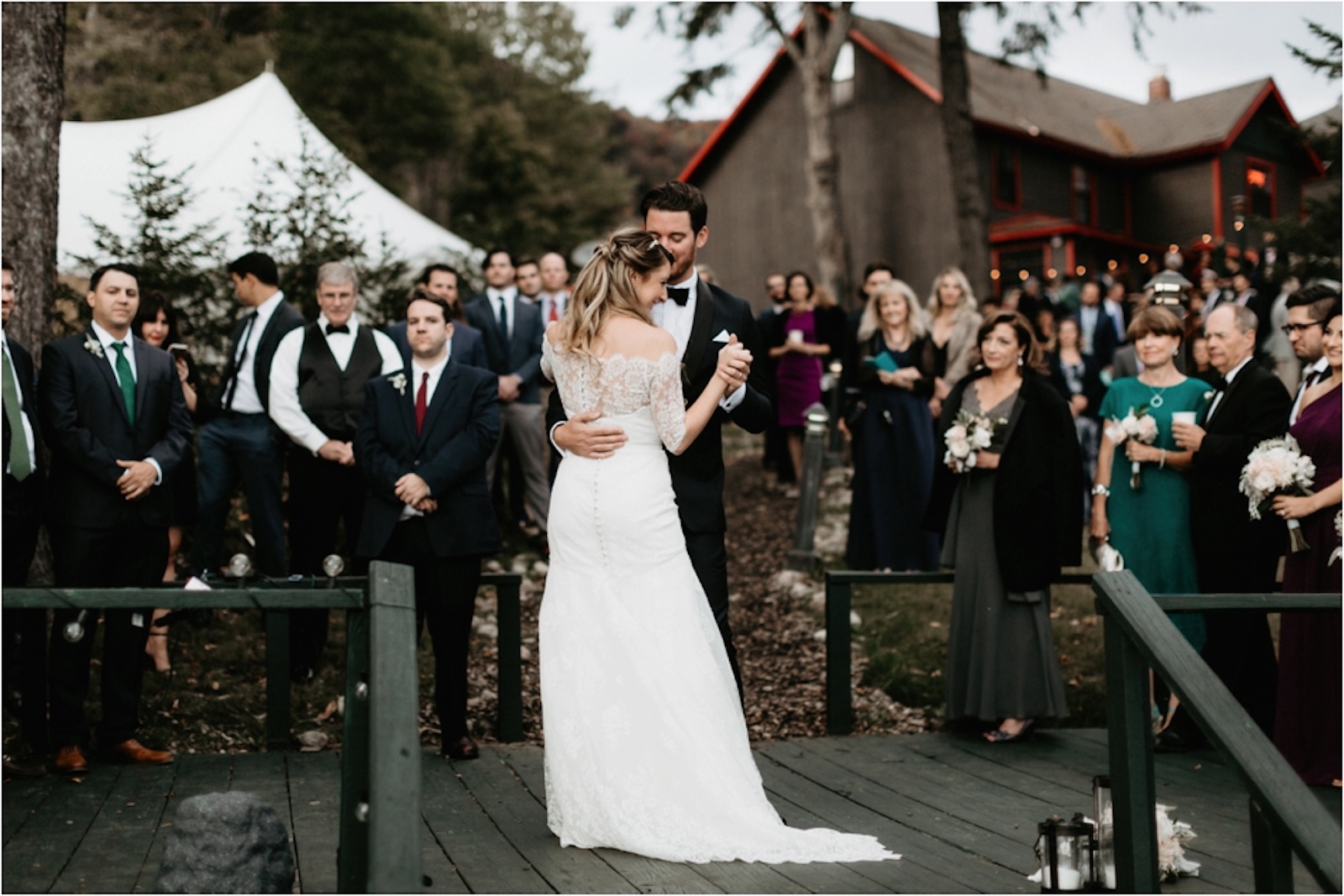 A bride and groom share their first dance on an outdoor wooden platform, surrounded by wedding guests watching and standing in formal attire.