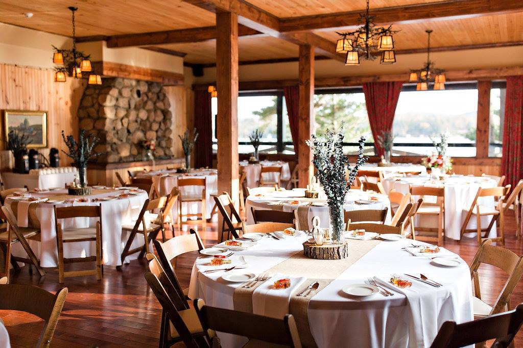 A rustic dining room with round tables set for an event, white tablecloths, wooden chairs, and centerpieces, with large windows and a stone fireplace in the background.