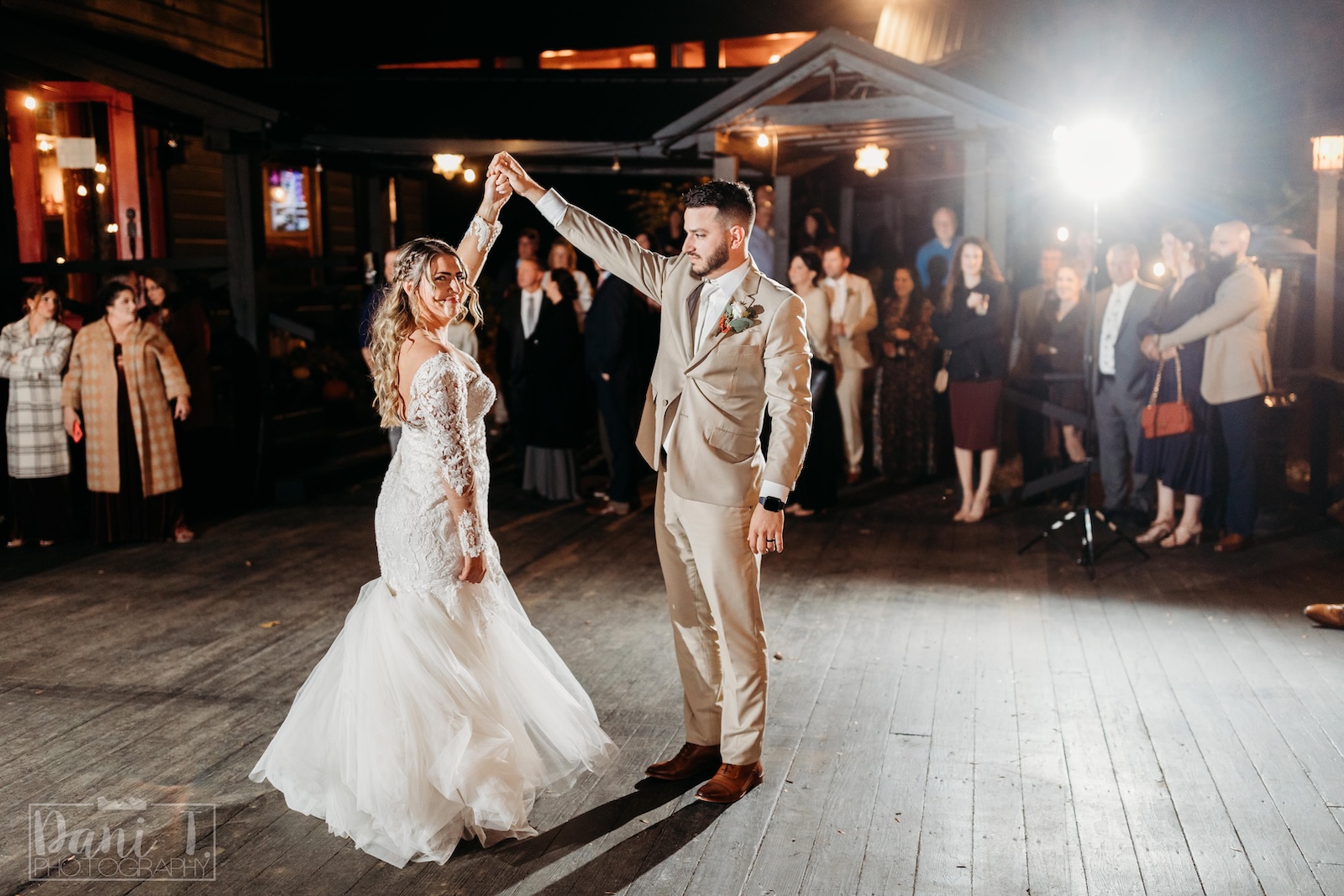 A bride and groom dance together on an outdoor wooden floor at night, surrounded by guests watching in the background.