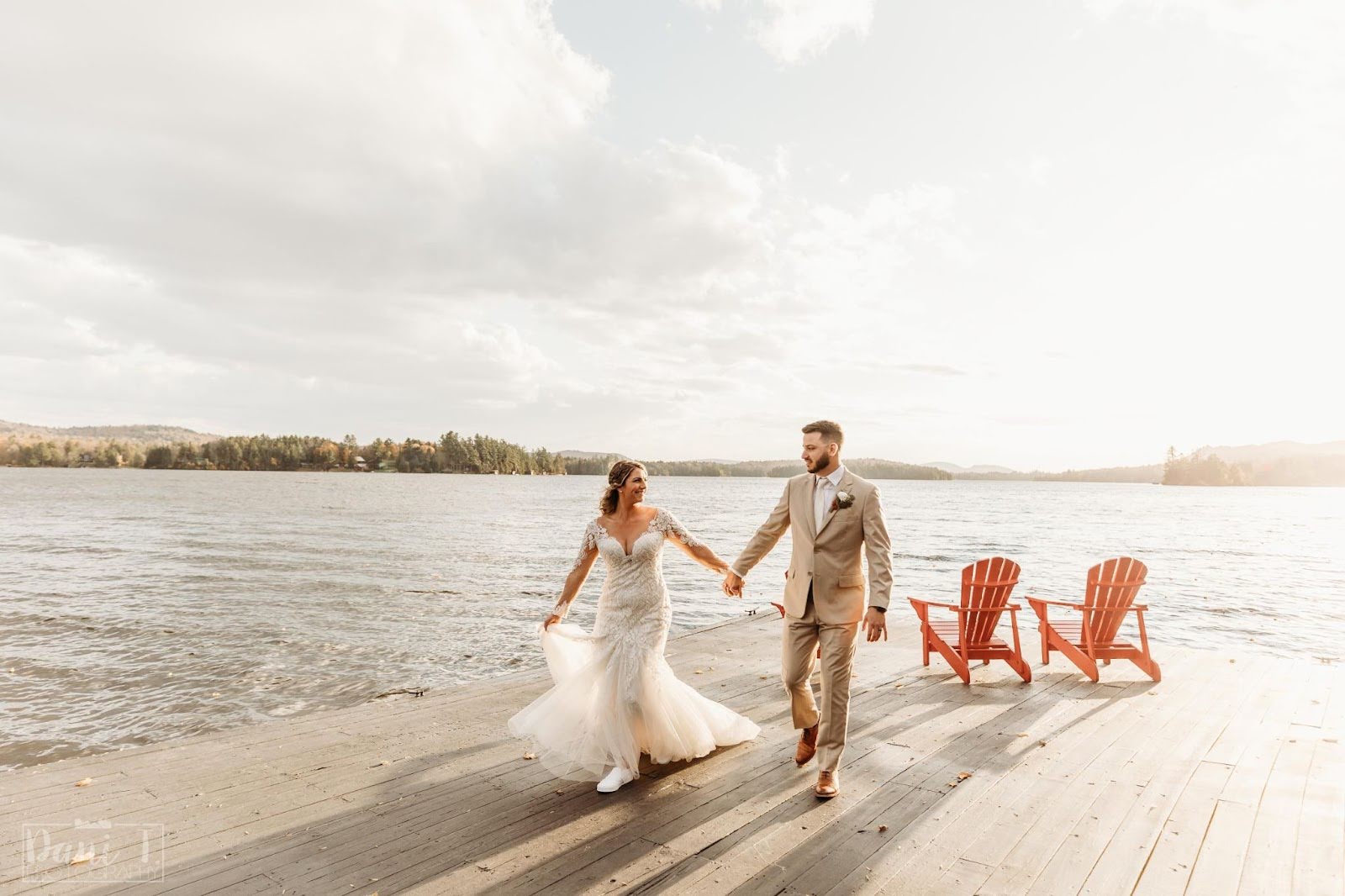 A bride and groom walk hand in hand on a wooden dock by a lake, with two red Adirondack chairs nearby and a cloudy sky overhead.