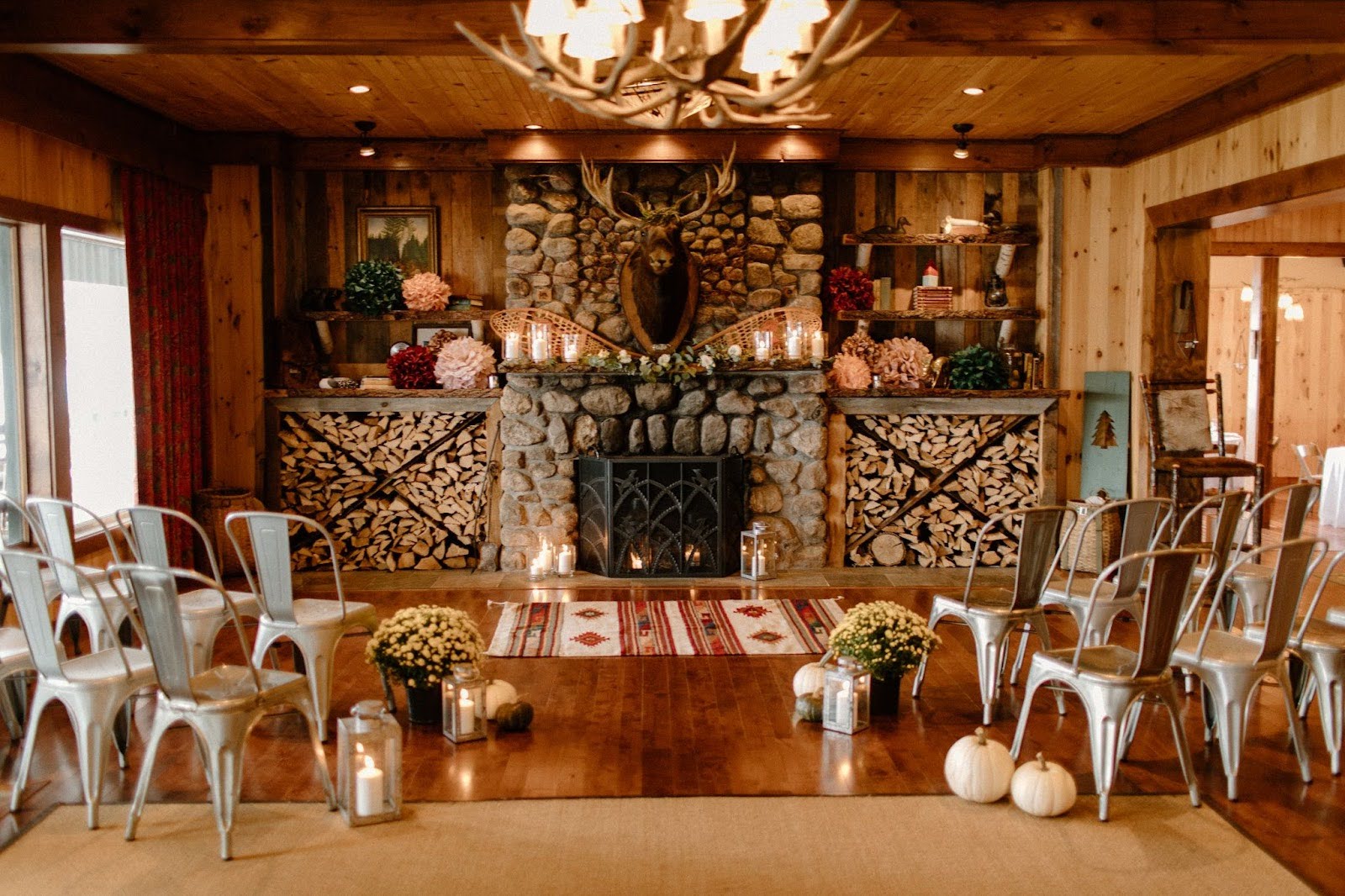 A rustic indoor ceremony setup with metal chairs facing a stone fireplace, decorated with flowers, candles, firewood, and pumpkins on a wooden floor.