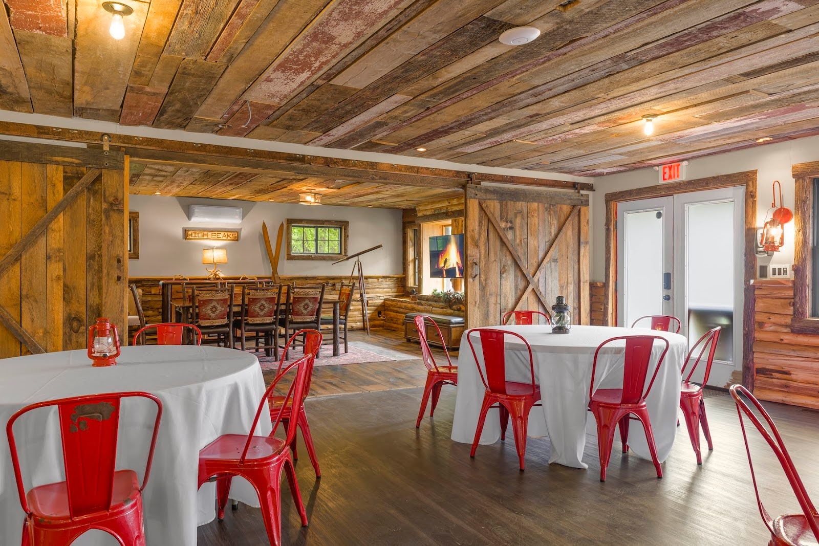 Rustic dining room with wooden walls and ceiling, round tables covered with white cloths, and red metal chairs; a wooden dining area is visible in the background.
