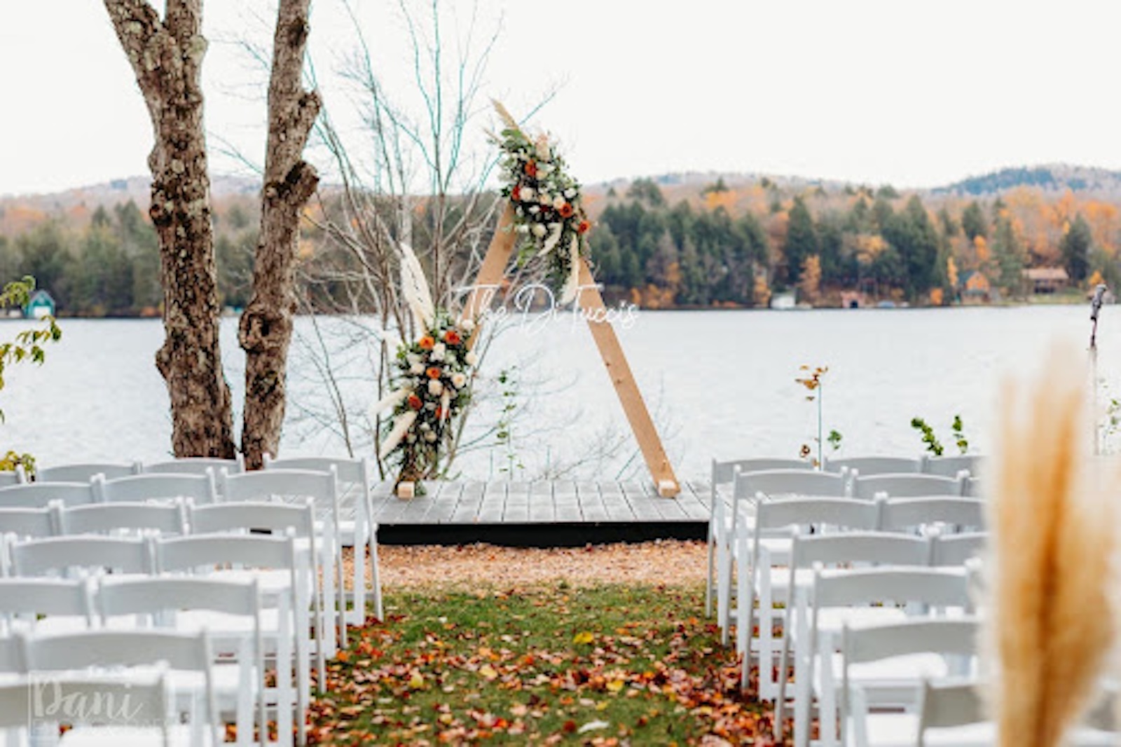 Rows of white chairs face a wooden wedding arch decorated with flowers, set on a dock by a lake with autumn trees in the background.