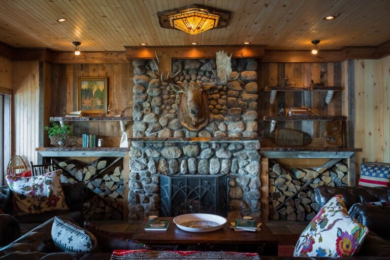 A rustic living room features a stone fireplace with a mounted moose head, wood paneling, shelves, firewood, leather chairs, and decorative pillows.