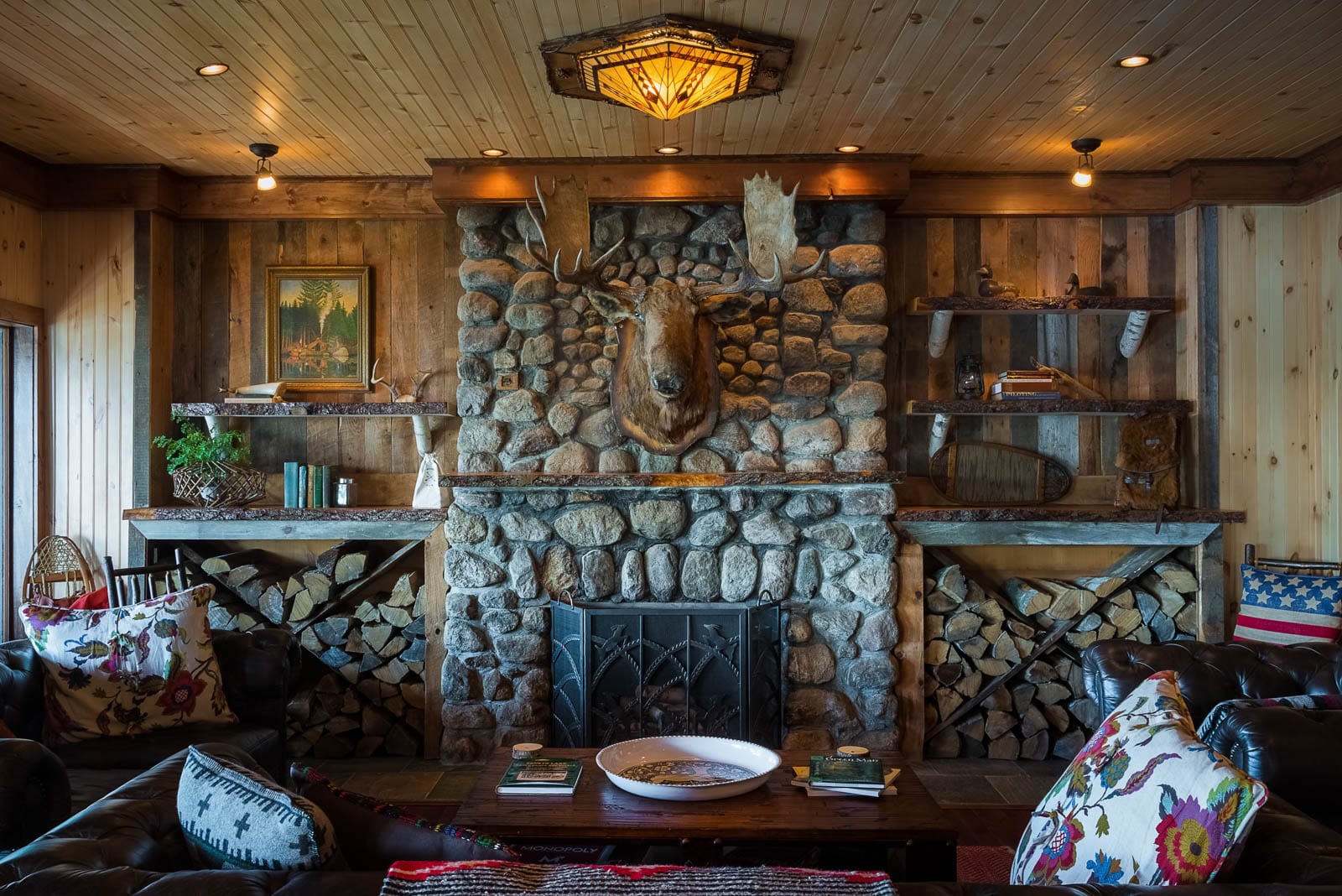 A rustic living room features a stone fireplace with a mounted moose head, wood paneling, shelves, firewood, leather chairs, and decorative pillows.