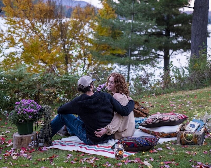Two people sit closely together on a blanket outdoors, surrounded by autumn leaves, flowers, and pillows, with trees and a lake in the background.