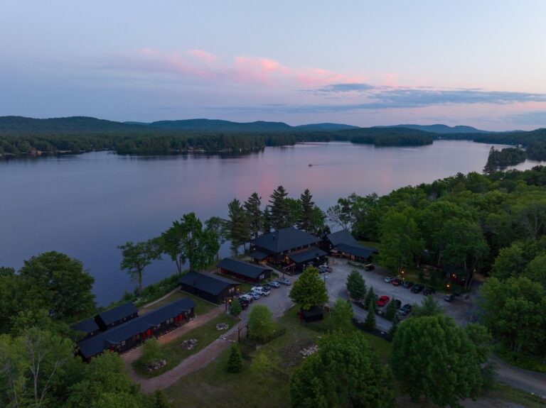 Aerial view of a lakeside lodge surrounded by trees at sunset, with calm water and rolling hills in the background.